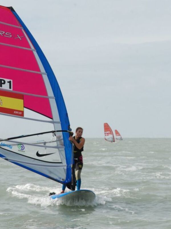 Un windsurfista en acción con vela roja y azul, navegando en aguas abiertas bajo un cielo nublado.