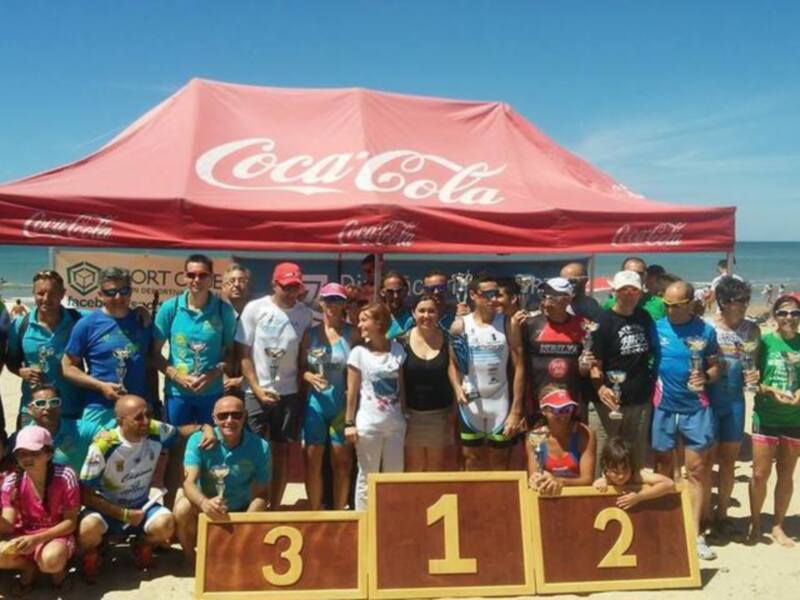 Equipo ganador de la carrera de triatlón en la playa, con premios y bandera de Coca-Cola en el fondo.