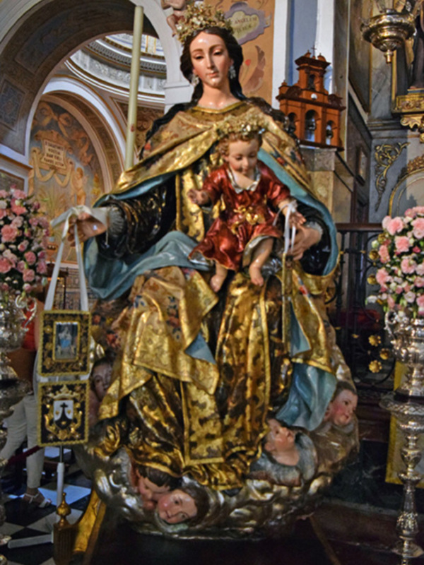 Imponente escultura de la Virgen María con el Niño Jesús, rodeada de flores y adornos dorados en un ambiente religioso.