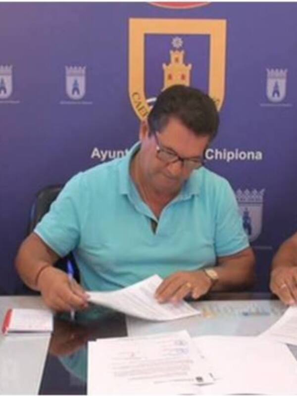 Tres personas firmando documentos en una mesa con un fondo de la bandera de Chipiona. El texto 'Ayuntamiento de Chipiona' se puede leer en el fondo.
