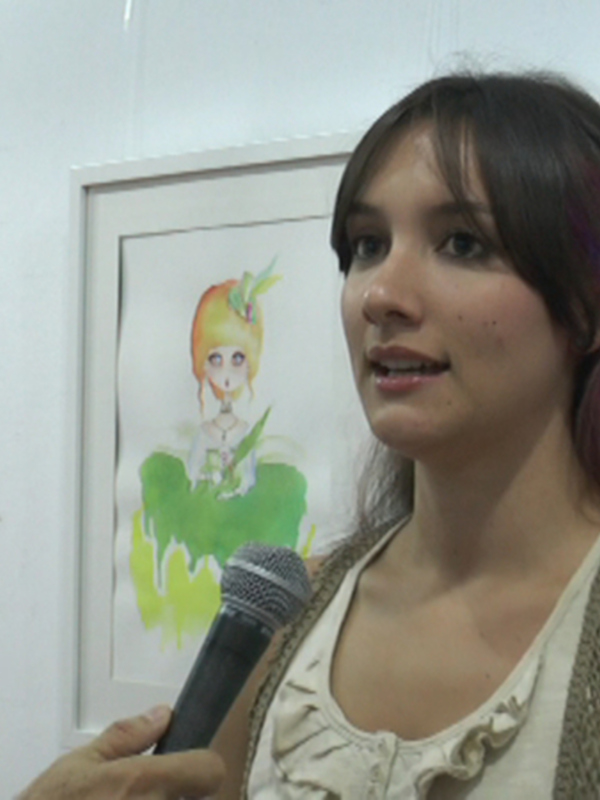 Una mujer con cabello largo y colorido habla en un micrófono frente a varias pinturas de personajes animados.