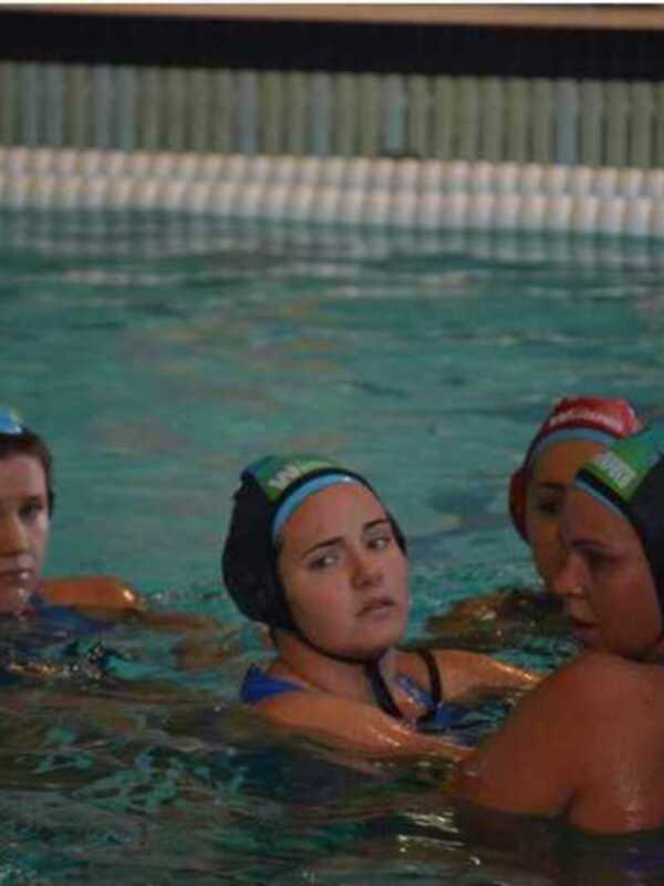 Jugadoras de waterpolo en una piscina, con cascos de protección y una figura en el fondo.