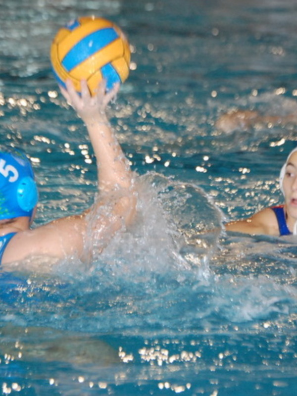 Jugadores de waterpolo en acción durante un partido, con un jugador en el centro intentando controlar el balón en el agua.