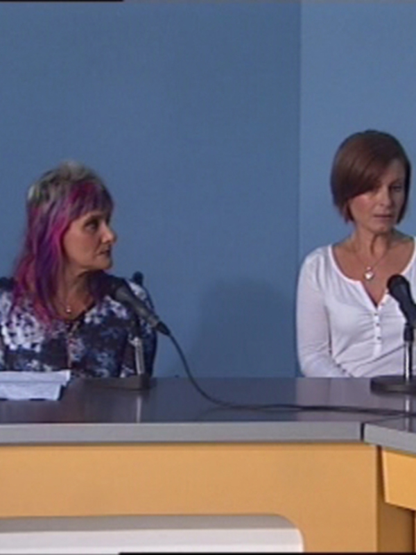 Dos personas se sientan frente a una pantalla azul en un entorno de conferencia o presentación. La persona izquierda tiene cabello rosa y usa una camisa con estampado floral, mientras que la derecha lleva un blusa blanca. Ambas tienen micrófonos en frente de ellas, y el escritorio tiene un portátil abierto.