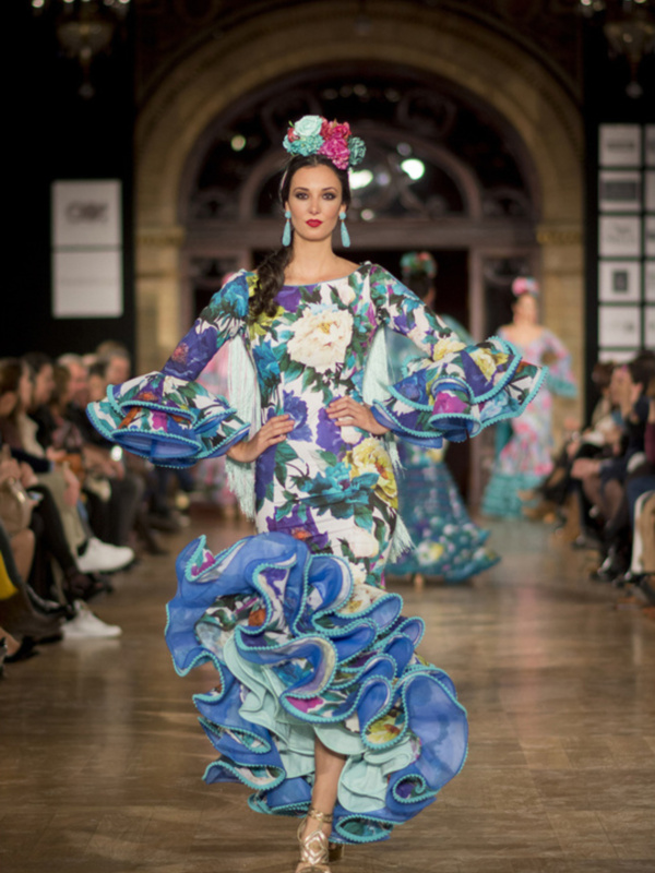 Una modelo desfilando en la pasarela de moda, con un vestido floral vibrante y una corona que resalta su belleza. La escena se centra en la moda, con detalles de color y diseño impresionantes.