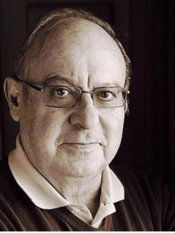 Retrato de un hombre con gafas y una expresión seria, vestido con una camisa de cuello alto y suéter oscuro, en un entorno interior con cortinas de fondo.