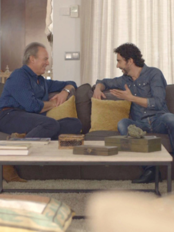 Dos hombres conversan en un sofá de salón con cojines amarillos, rodeados de decoración minimalista y natural.