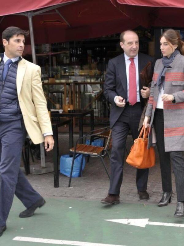 Tres personas caminan juntas por una calle urbana, una con abrigo y bolso naranja, otra con chaqueta beige y la tercera con traje y corbata roja.