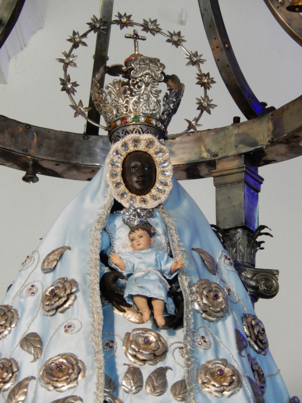 Imaginación de una Virgen María con un niño Jesús en sus brazos, vestida con una saya azul y adornada con flores doradas y cristales, sostenida por una estructura metálica con detalles ornamentales.