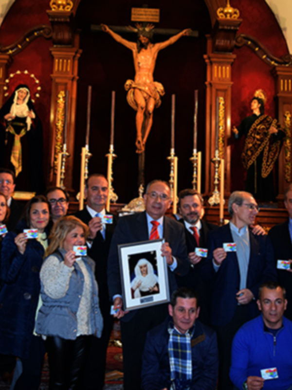 Una imagen de una iglesia con un altar central y varios personajes en el fondo. Los individuos están posando para una foto, con algunos sosteniendo tarjetas de identificación. La decoración incluye un crucifijo y estatuas religiosas en el fondo, iluminada por luces de araña.