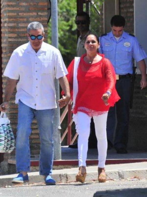 Una pareja camina juntos por una calle, sosteniendo las manos, con un guardia de seguridad detrás.