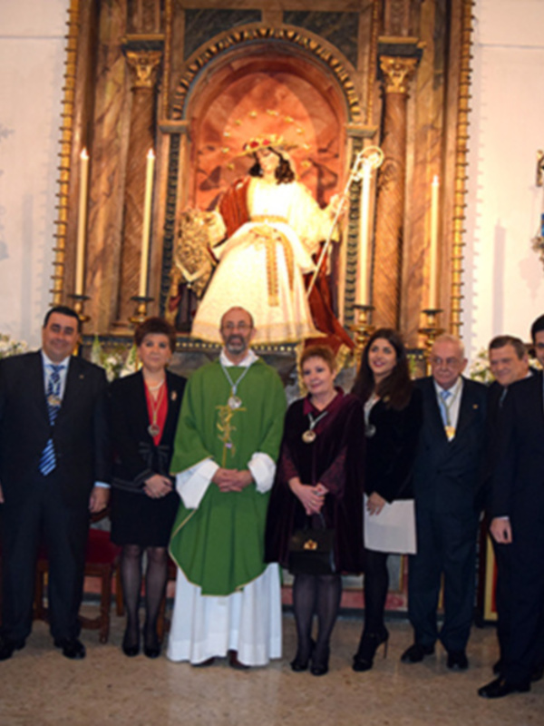 Una imagen de una ceremonia religiosa en un templo, con una estatua central y varias personas vestidas formalmente. La decoración incluye velas, una bandera y elementos arquitectónicos ornamentados.