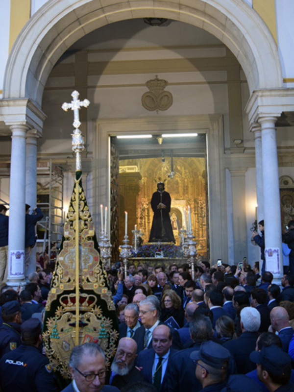 Una procesión religiosa en un templo con una cruz dorada y un altar en el fondo, rodeada por una multitud de personas vestidas formalmente.