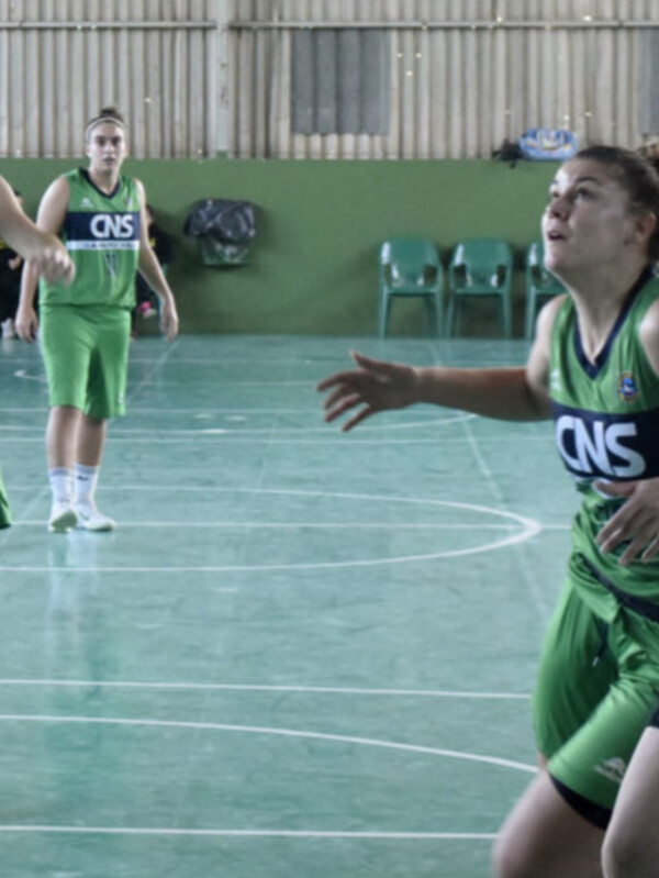 Partido de baloncesto femenino en un gimnasio, con jugadoras de dos equipos, uno con uniforme verde y otro con negro y amarillo, en una cancha de baloncesto interior.