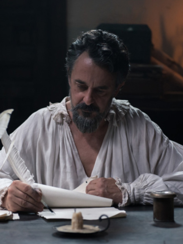 Hombre en vestimenta medieval escribiendo con pluma en una mesa con candelabro y libro antiguo.