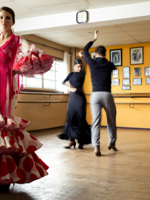 Dancers in a dance studio, with a woman in a vibrant red and white flamenco dress and a man in a black top and grey pants practicing together.