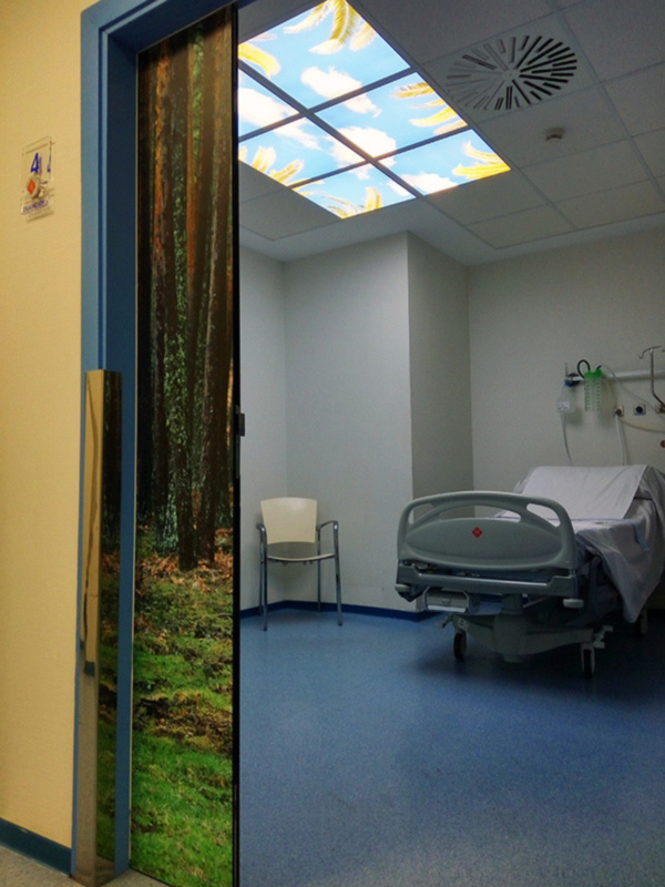Interior de un hospital con cama de hospitalización, silla y techo con iluminación de diseño de bosque.
