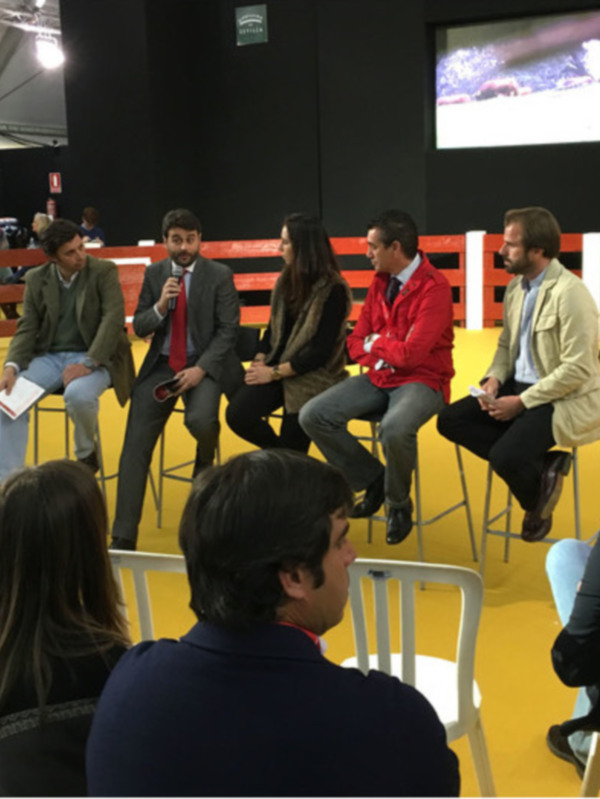 Discusión en un evento sobre ganado, con participantes sentados en un escenario rodeado de audiencia y un toro en el fondo.