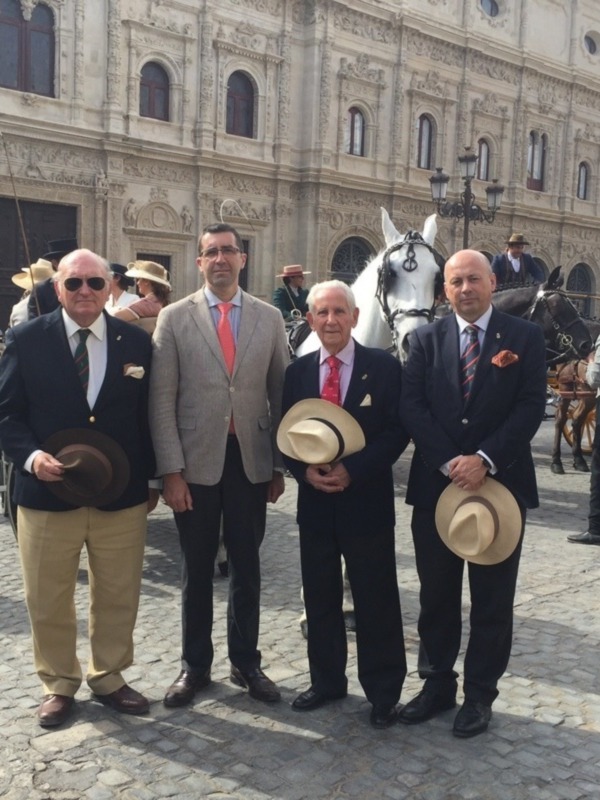 Cuatro hombres vestidos formalmente posan frente a un edificio histórico con un caballo y carruaje en el fondo.