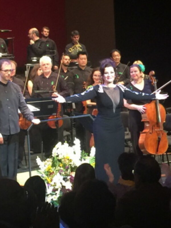 Una orquesta se presenta en un escenario con un solista en el centro, mientras el público disfruta de la música.