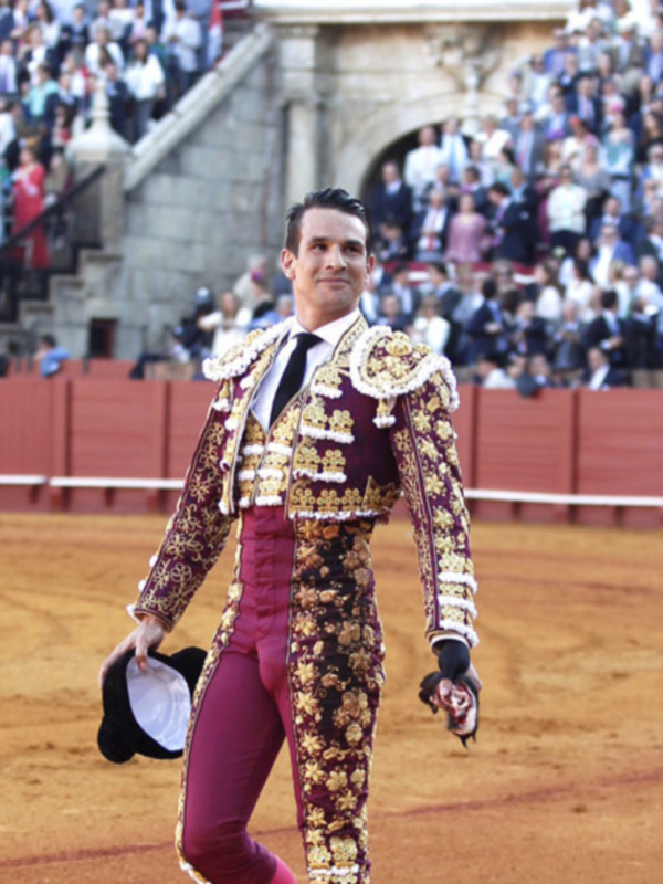 Un torero en traje de luces camina por la plaza de toros, saludando a la multitud en el fondo.