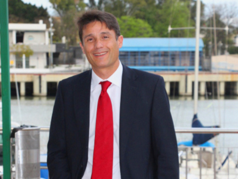 Elegante hombre en traje y corbata roja, posando frente a un mar tranquilo con embarcaciones y estructuras en el fondo.