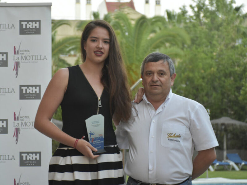 Una mujer sostiene un premio junto a un hombre, ambos posan frente a un fondo con logos de hotel y marcas comerciales.