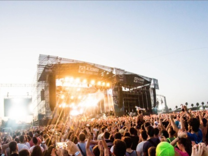 Una multitud de personas disfrutando de un concierto al aire libre en una tarde soleada. La plataforma central está iluminada con luces brillantes y el público se alza en un ambiente festivo.