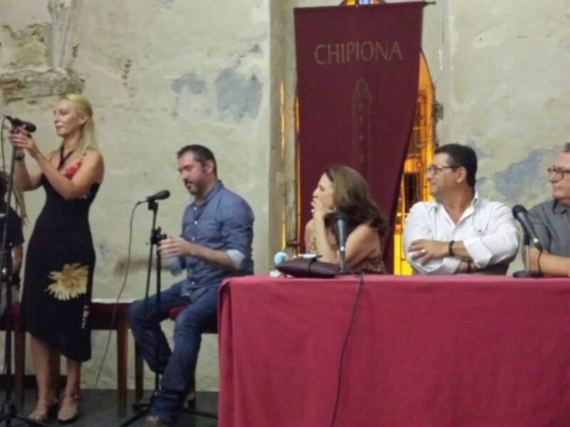 Una conferencia de prensa en un edificio histórico, con personas sentadas en una mesa y un panel de micros en el fondo. La bandera de Chipiona está visible en el fondo.
