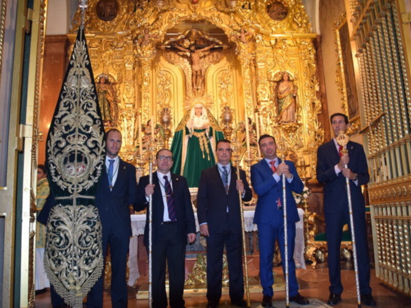 Cuatro hombres en trajes de negocios posan con una bandera frente a una imagen religiosa dorada en una iglesia.
