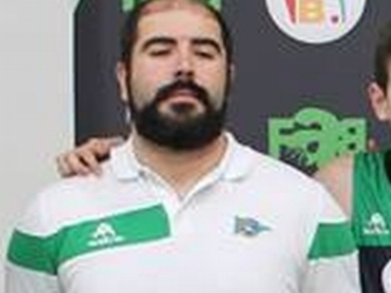 Fotografía de un hombre con barba y una camiseta blanca y verde, posando con una persona en el fondo.