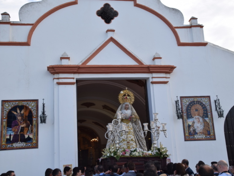 Procesión de la Virgen de la Caridad en una iglesia blanca con detalles decorativos y una multitud en la plaza.