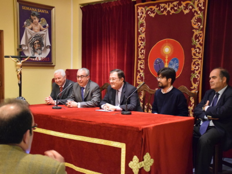 Conferencia en el marco de la Semana Santa, con figuras religiosas y decoración festiva en el fondo.