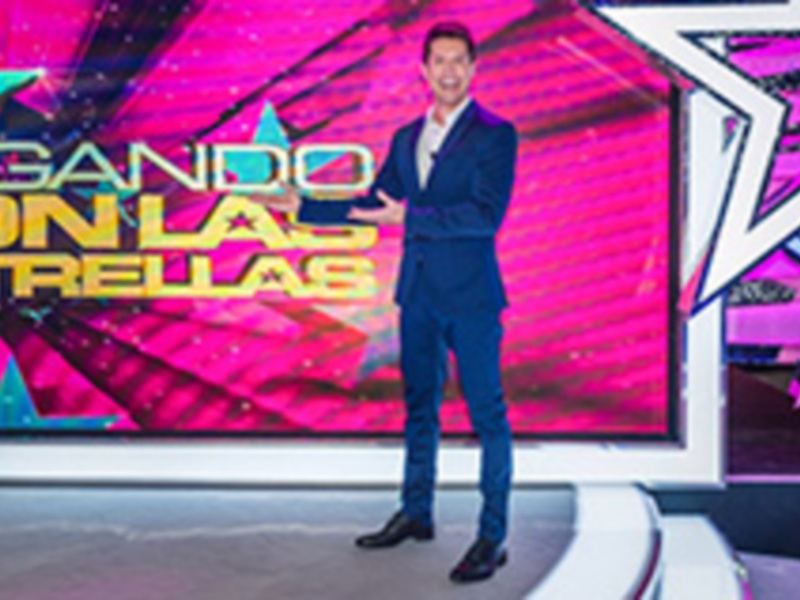 Presentador en escenario con fondo vibrante y texto 'JUGANDO CON LA ESTRELLA'