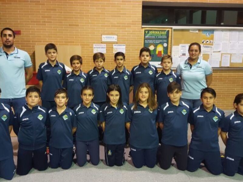 Un grupo de niños y niñas en uniforme azul se posan frente a una pared con carteles. Al fondo, un hombre en camiseta azul se inclina para tomar la foto. La escena parece ser una reunión o sesión de fotos escolar en un ambiente interior, posiblemente una escuela.