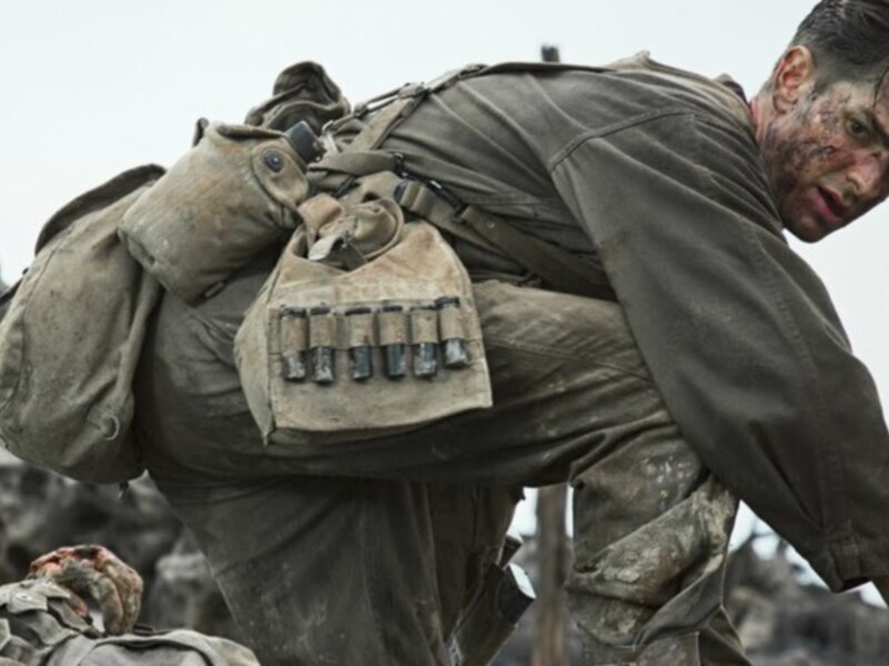 Un soldado en uniforme militar, cargando una mochila llena de balas. El fondo es despejado y gris, sugiriendo un entorno militar o de combate.