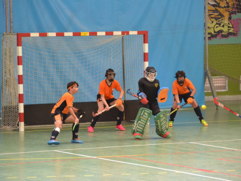 Jugadores de hockey en acción en un campo interior, con un portero en acción y otros jugadores en espera de la pelota.