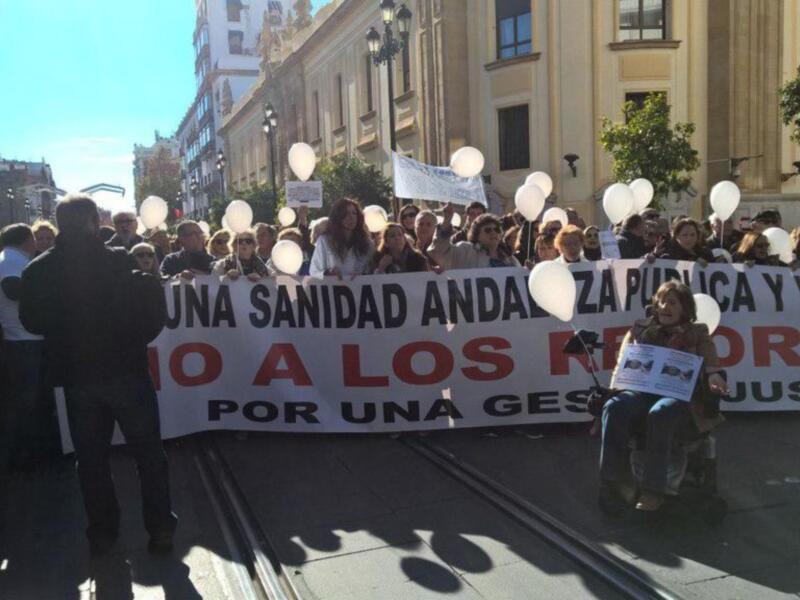 Una multitud de personas se manifiesta en una calle con pancartas que dicen "Una sanidad andaluza pública y única. No a los cortes. Por una gestión justa." El ambiente es de protesta y solidaridad, con un cielo azul claro.