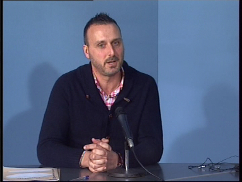 Hombre hablando en un evento con una camisa a cuadros y un suéter oscuro, frente a un micrófono.