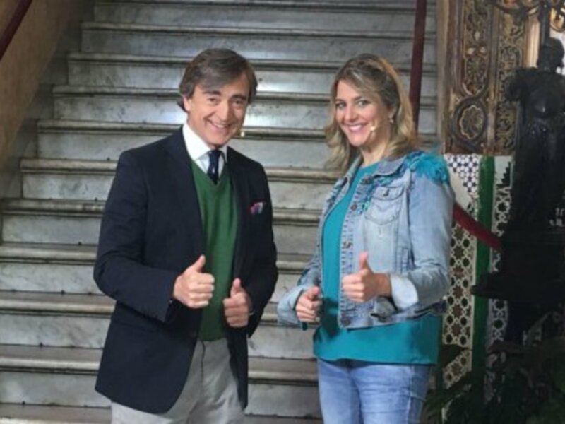 Dos personas sonrientes con gesto de aprobación, uno con chaqueta y corbata y el otro con chaqueta vaquera. Fondo de escaleras y decoración clásica.