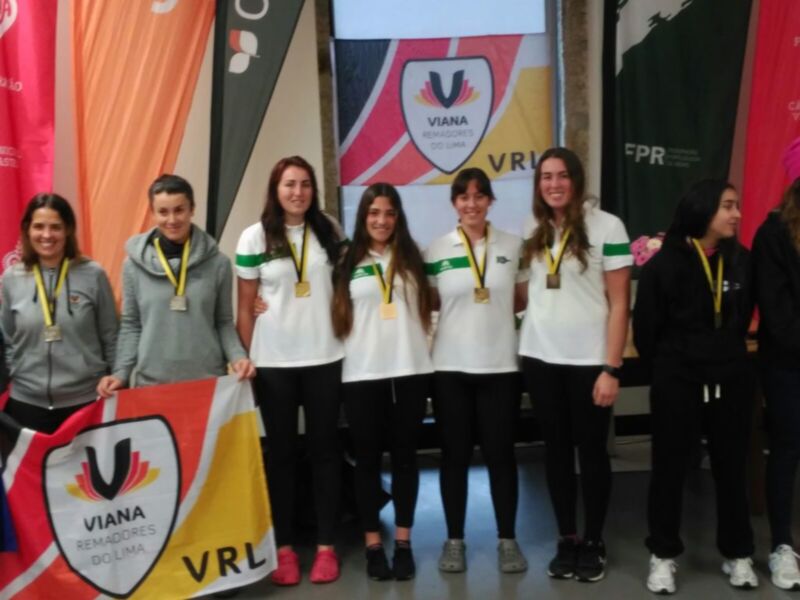 Equipo femenino de remo posando con sus medallas y la bandera de Viana Remadores del Lima en un evento deportivo.