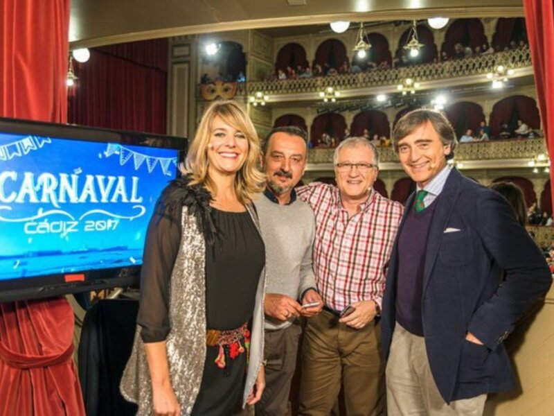 En un escenario teatral, tres personas son entrevistadas en el centro. A la izquierda, una mujer con cabello largo y vestida de negro y plateado sonríe ampliamente. A la derecha, un hombre con barba y camisa roja sonríe mientras sostiene una tableta. En el fondo, una pantalla grande muestra "Carnaval" y "CCAPZ 2017". La escena está iluminada con luces de teatro y el telón rojo del escenario se puede ver a la izquierda.