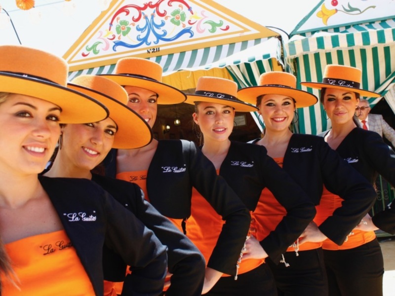 Cuatro mujeres vestidas con trajes de color naranja y negro, con sombreros amarillos, posan frente a un toldo festivo.