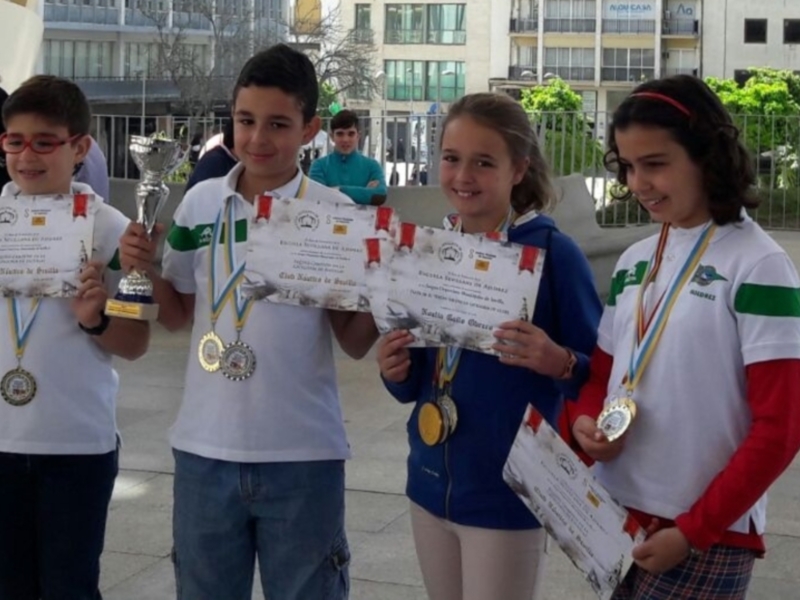 Tres niños sonrientes sostienen trofeos y diplomas, con un edificio de oficinas en el fondo.