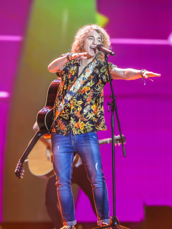Artista con guitarra y camisa floral en escenario con luces de fondo vibrantes.