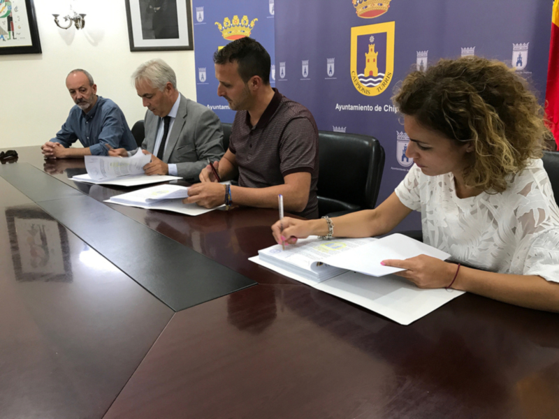 En una sala de conferencias, tres personas se sientan alrededor de una mesa. Dos hombres y una mujer están firmando documentos, con el hombre en medio escribiendo mientras los otros dos observan. La mujer a la derecha lleva una pulsera y un reloj, mientras que el hombre en medio tiene una corbata. En la pared detrás de ellos, hay un panel con el escudo del Ayuntamiento de Chicl.