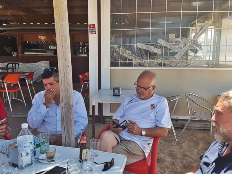 Tres hombres sentados en una terraza, conversando y tomando bebidas. Detalle de una botella de agua natural en el primer plano. El ambiente es casual y relajado, con sillas y mesas dispuestas para el descanso.