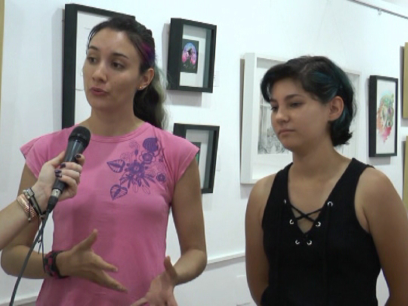 Dos jóvenes conversan en una galería de arte, con cuadros colgados en el fondo. La imagen captura un momento de diálogo entre ellas, enfocándose en su expresión y gesto.