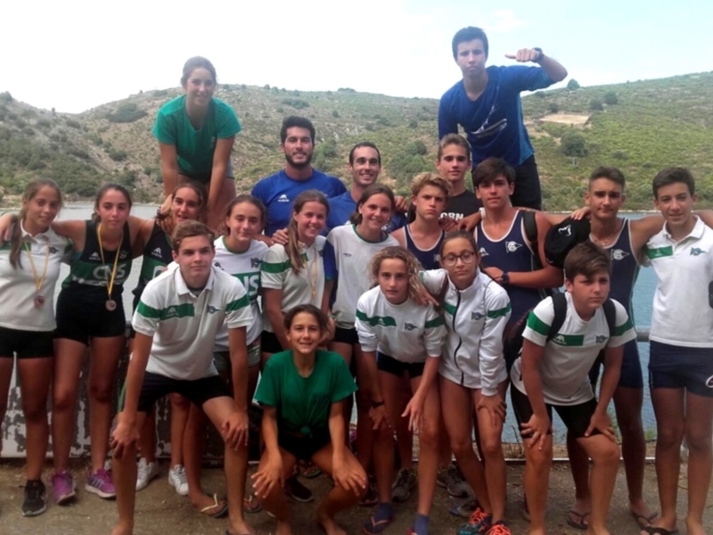 Una escuela de atletismo posa para una foto en un entorno natural con un lago en el fondo. Los atletas están vestidos con uniformes deportivos, y algunos llevan medallas de oro, lo que sugiere que han participado en una competición o evento deportivo.