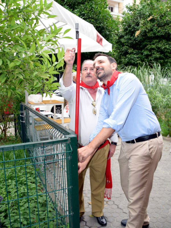 Dos hombres interactúan con un poste rojo en un entorno verde y urbano.